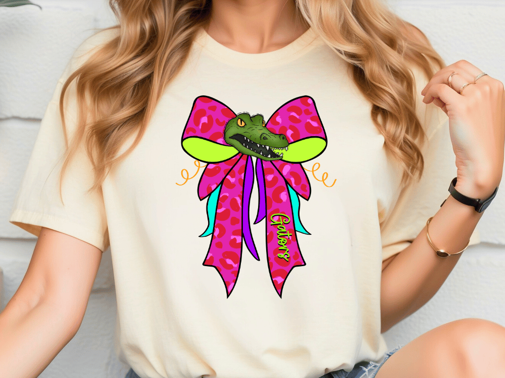 A woman wearing a white t-shirt with a colorful graphic design featuring a green alligator wearing a pink bow.