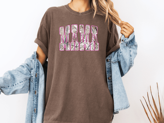 A woman wearing a brown t-shirt with the word "MAMA" printed on it, along with a denim jacket, stands against a white background.