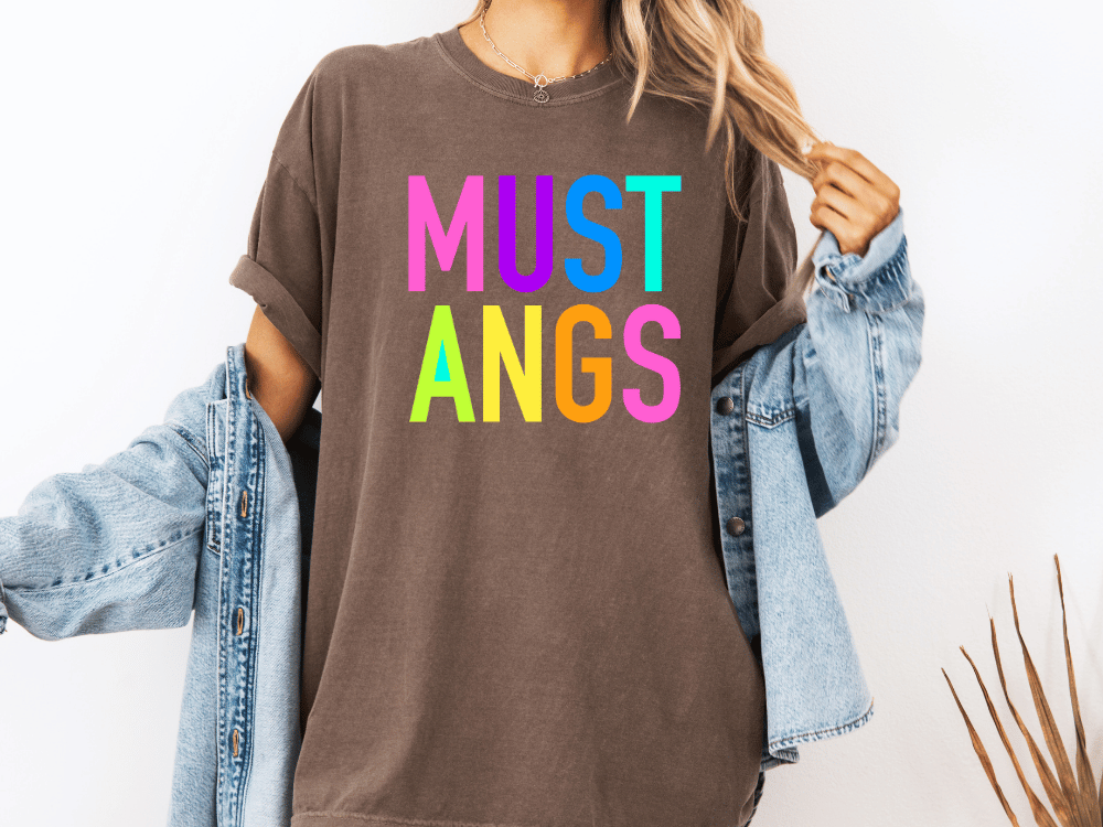 A person wearing a brown t-shirt with the text "MUST ANGS" in colorful letters, along with a denim jacket, stands against a white background.