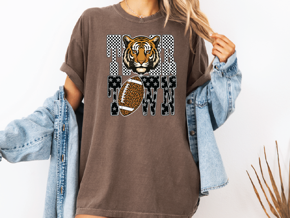 A woman wearing a brown t-shirt with a tiger graphic and a football, along with a denim jacket, stands against a white background.