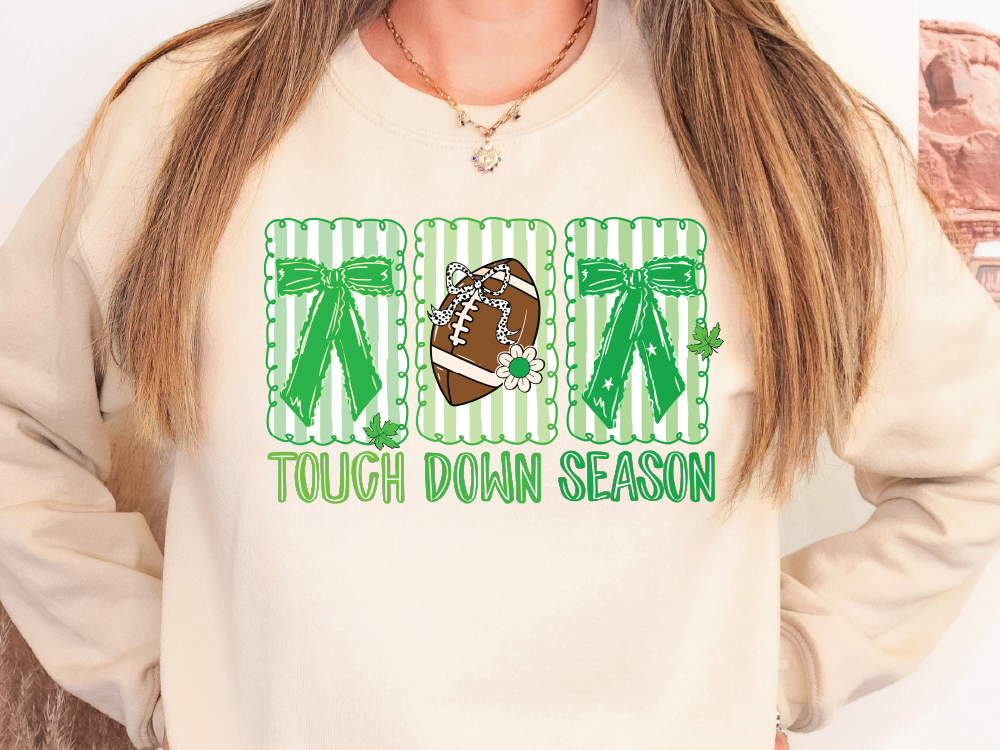 A woman wearing a white sweatshirt with a graphic design featuring a football, green bows, and the text "TOUCH DOWN SEASON".