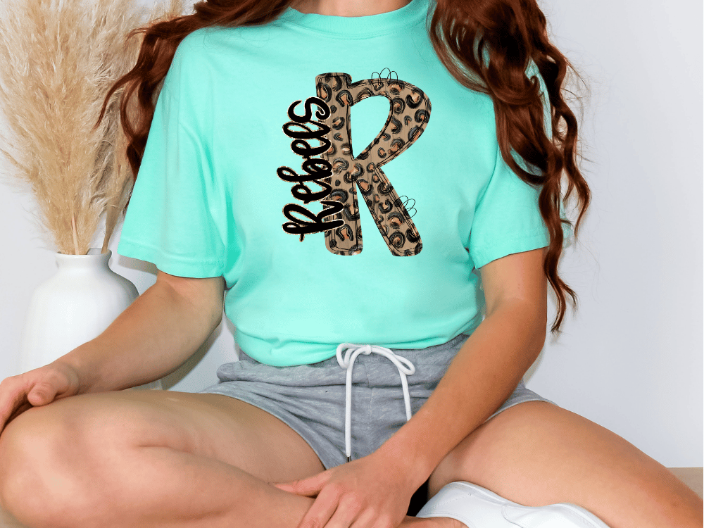A person wearing a mint green t-shirt with a leopard print design and gray shorts is sitting on the floor.