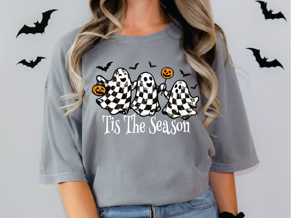 A woman wearing a gray t-shirt with a graphic design featuring three ghostly figures and the text "Tis The Season" printed on it.