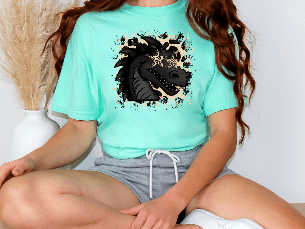 A woman wearing a turquoise t-shirt with a graphic of a stylized dragon or mythical creature on it, sitting cross-legged on the floor.