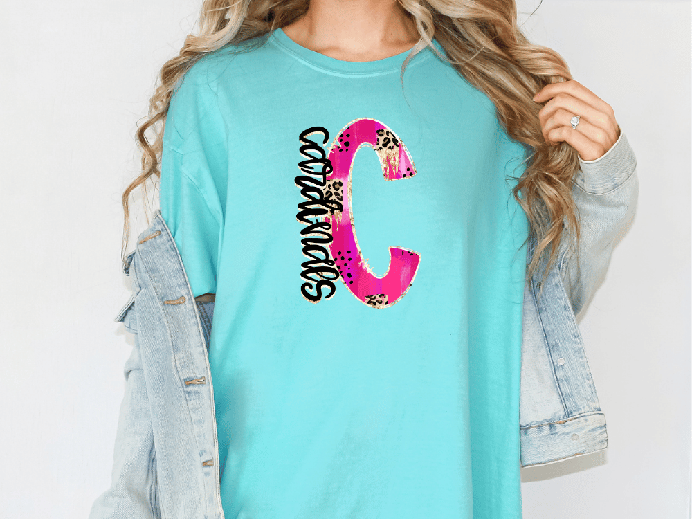 A woman wearing a turquoise t-shirt with a large pink and black letter 'C' printed on it, along with a denim jacket.
