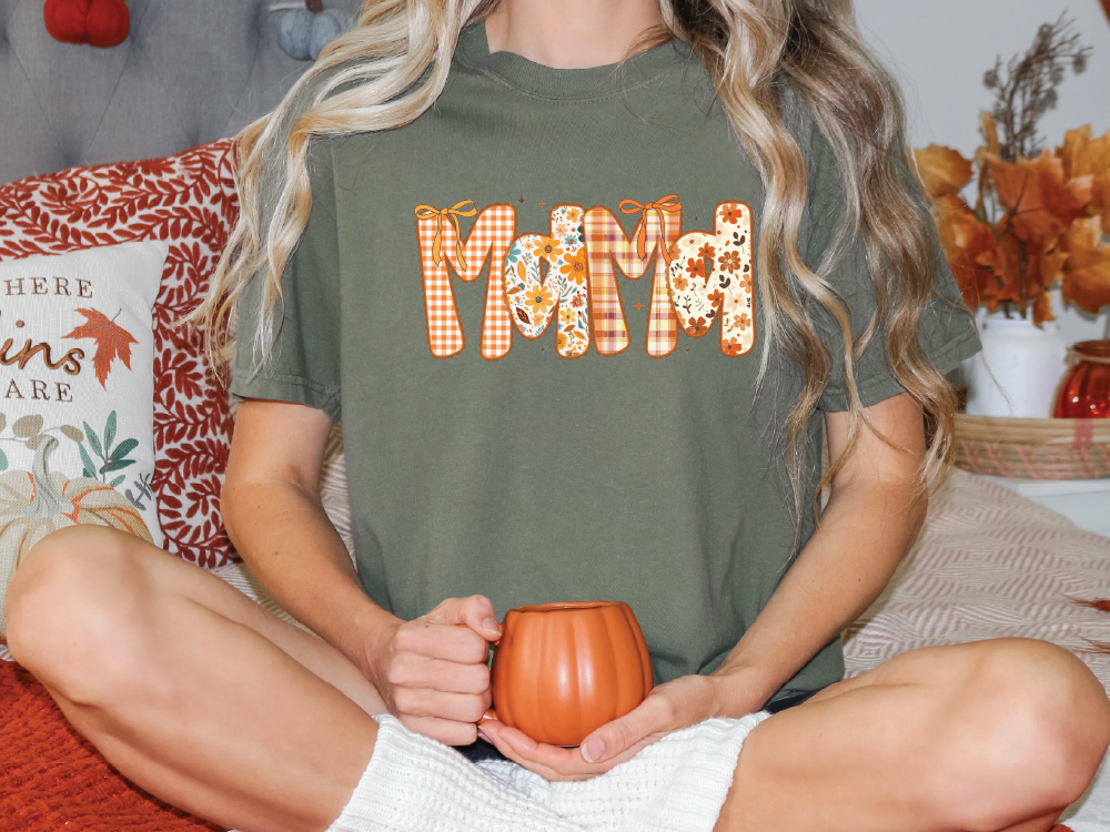 A woman wearing a green t-shirt with the word "Mama" printed on it, sitting on a bed with a pumpkin-themed pillow and holding a pumpkin-shaped mug.