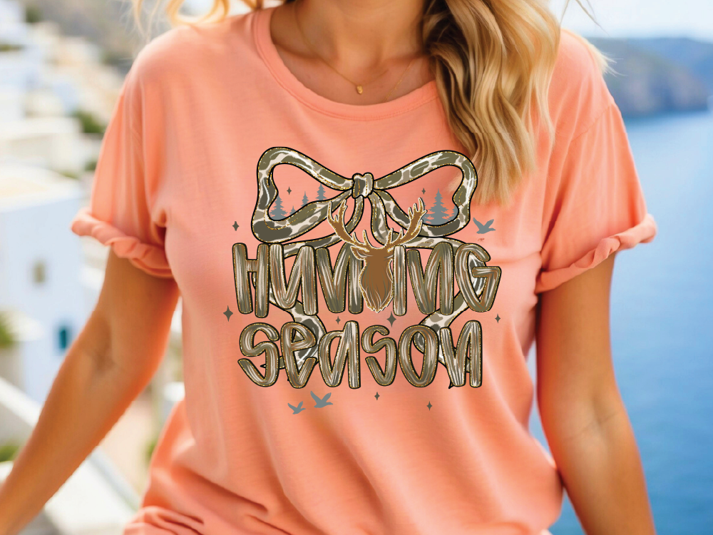 A woman wearing a peach-colored t-shirt with a graphic design featuring a bow and the text "HUMMING SEASON" printed on it.