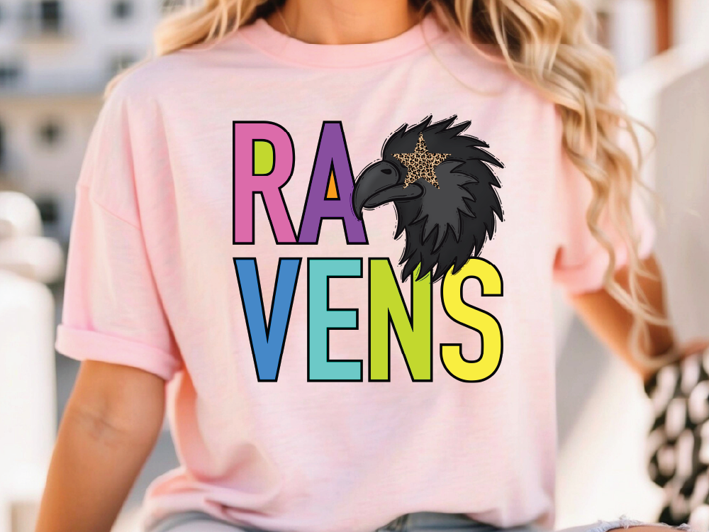 A woman wearing a pink t-shirt with a colorful graphic design featuring a stylized bird or bird-like figure and the word "RAVENS" written below it.