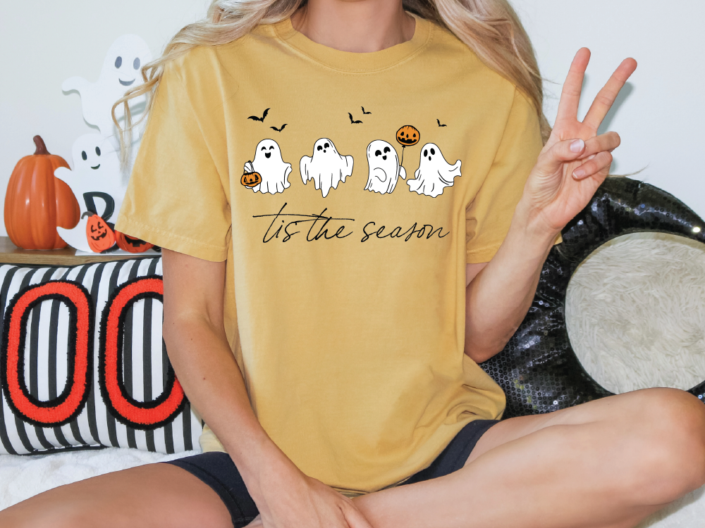 A person wearing a yellow t-shirt with a graphic of ghost characters and the text "Tis the season" is sitting on the floor.