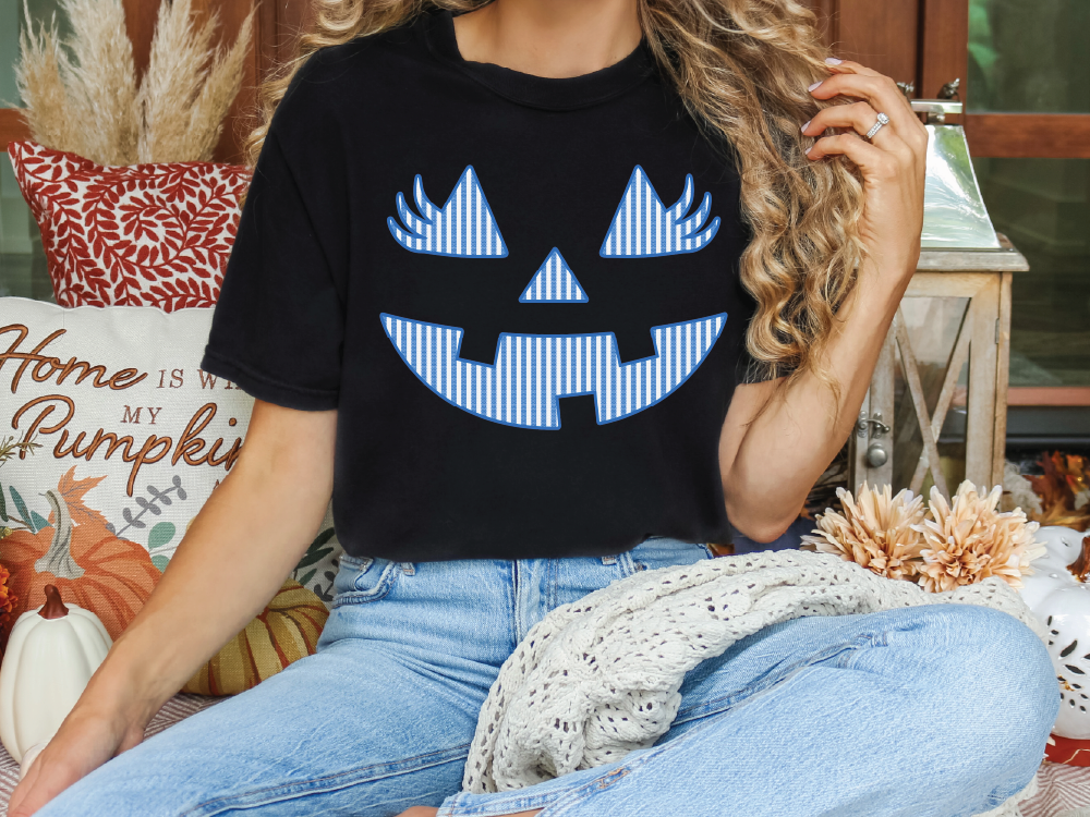 A woman wearing a black t-shirt with a blue jack-o-lantern design is sitting on a couch surrounded by fall decorations.