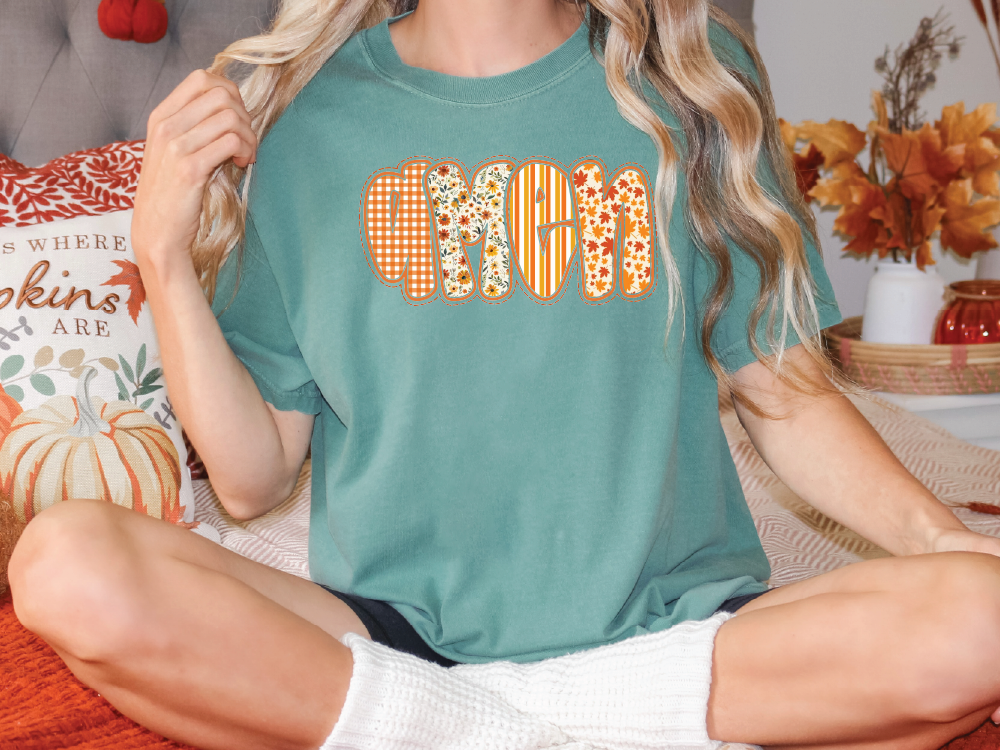 A woman wearing a teal t-shirt with the word "MOM" printed on it, sitting on a bed with autumn-themed pillows and decorations.