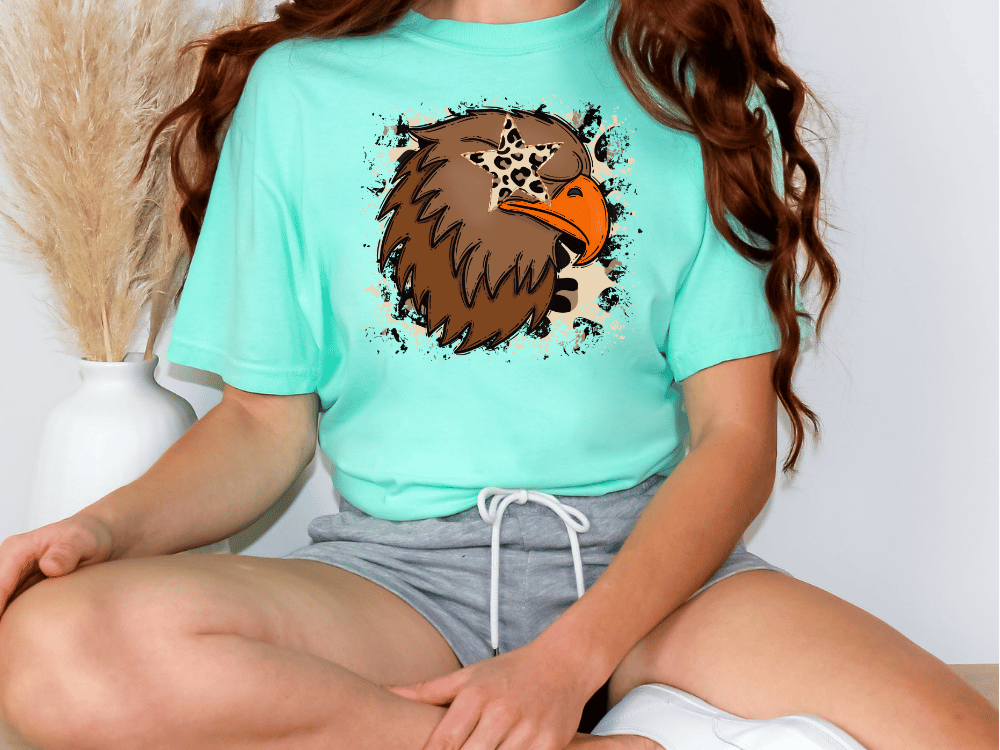 A person wearing a turquoise t-shirt with a graphic of a brown eagle and a star on it, sitting on the floor.