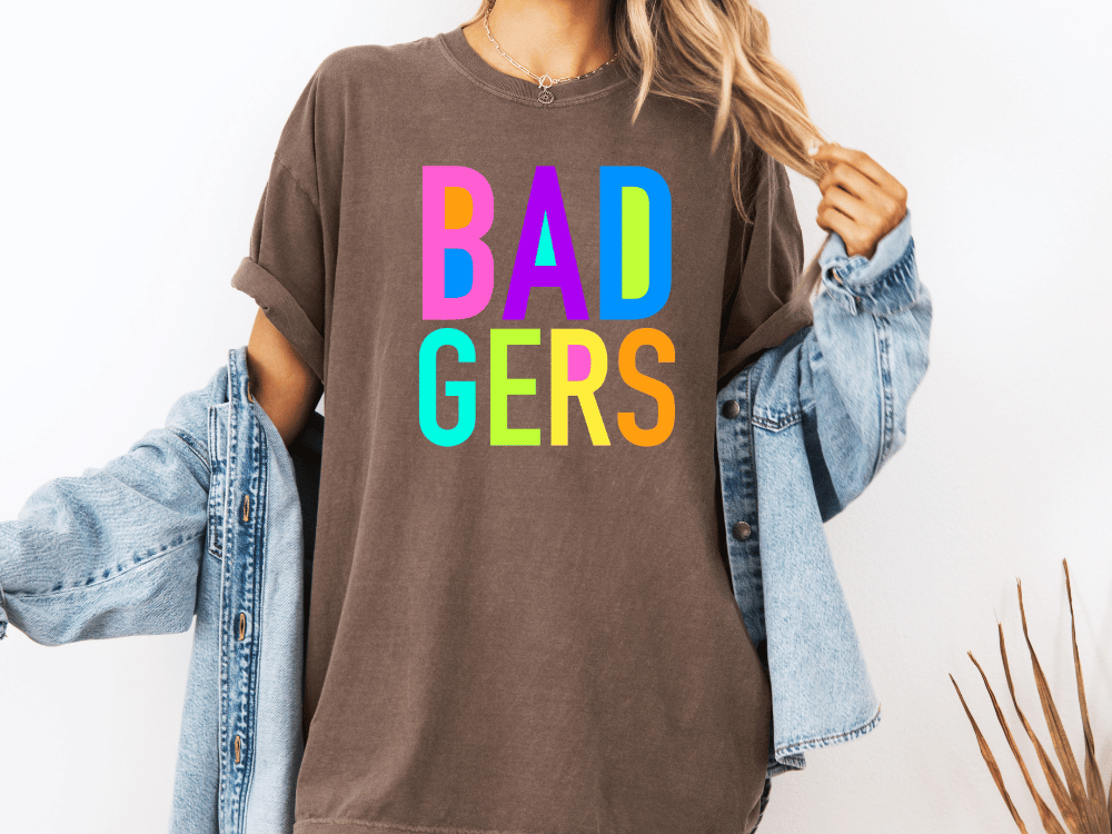 A person wearing a brown t-shirt with the text "BAD GERS" in colorful letters, paired with a denim jacket.