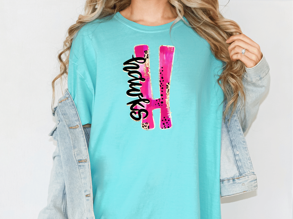 A woman wearing a turquoise t-shirt with a pink and black design on it, and a denim jacket.