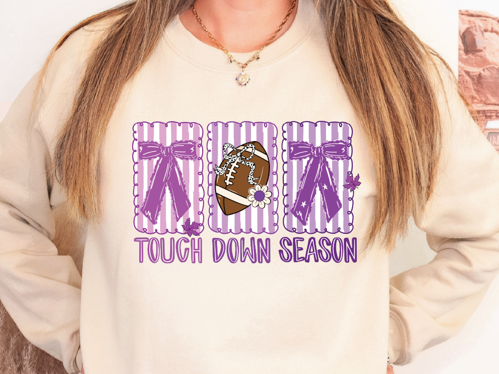 A woman wearing a white sweatshirt with a graphic design featuring a football and the text "TOUGH DOWN SEASON" printed on it.