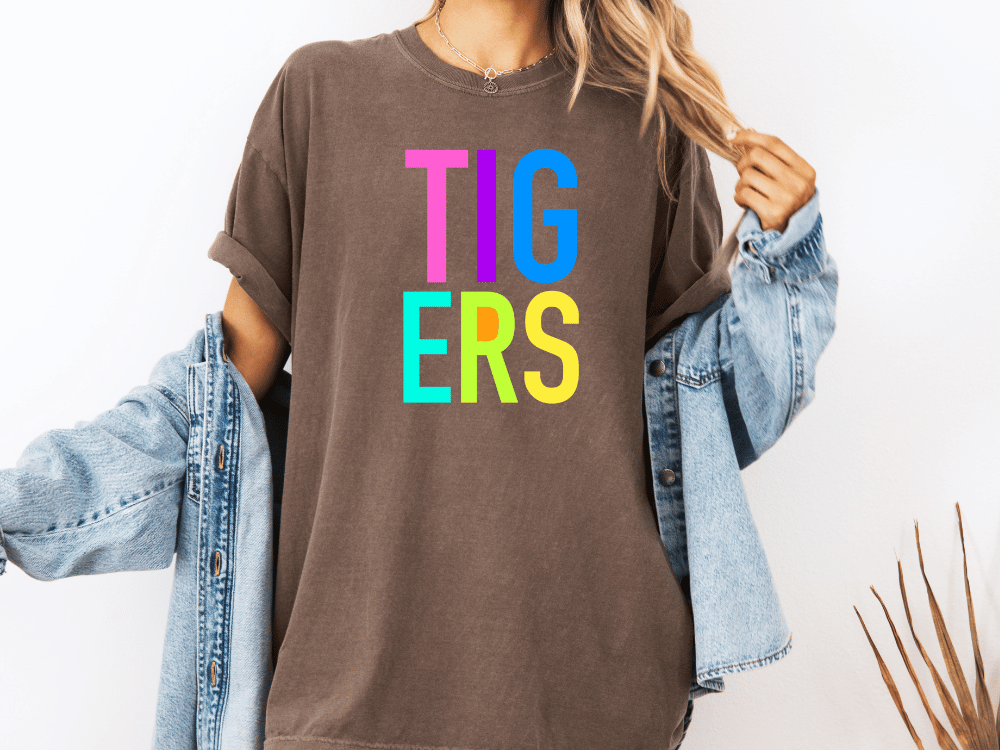 A person wearing a brown t-shirt with the word "TIGERS" in colorful letters, paired with a denim jacket, stands against a white background.