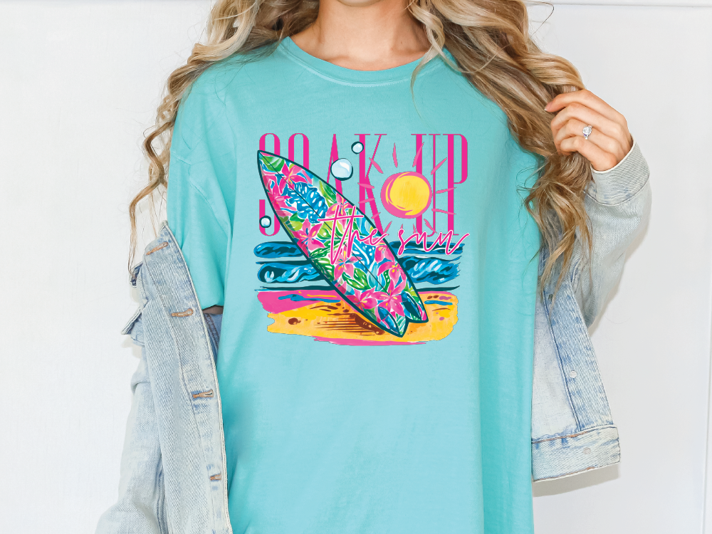 A woman wearing a turquoise t-shirt with a colorful graphic of a surfboard and beach scene.