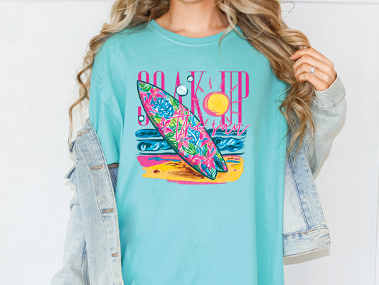 A woman wearing a turquoise t-shirt with a colorful graphic of a surfboard and beach scene.
