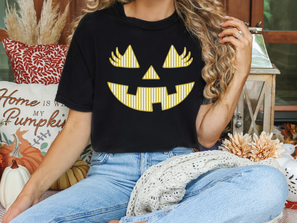 A woman wearing a black t-shirt with a pumpkin jack-o-lantern design is sitting on a couch surrounded by fall decorations.