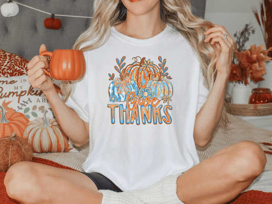 A woman sitting on a bed, holding a mug and wearing a white t-shirt with a pumpkin graphic and the text "Give Thanks".