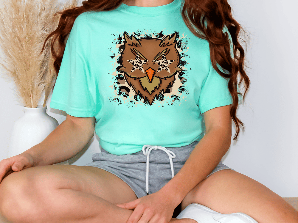 A person wearing a turquoise t-shirt with an owl graphic and gray shorts is sitting cross-legged on the floor.