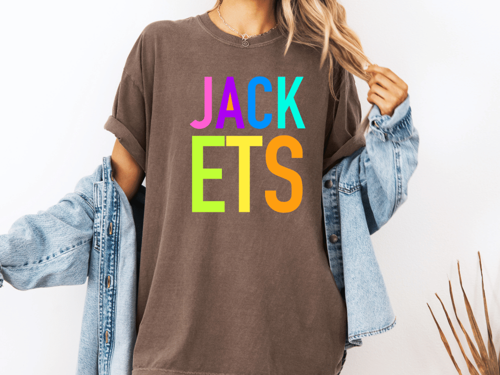 A person wearing a brown t-shirt with the text "JACK ETS" in colorful letters, along with a denim jacket, stands against a white background.