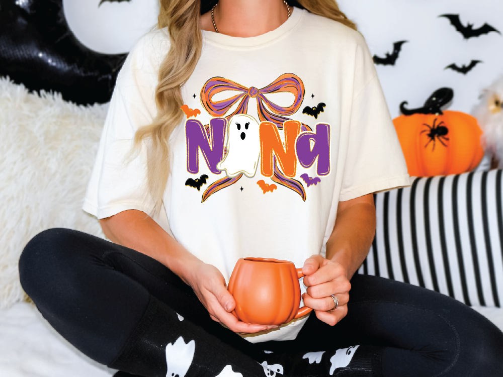 A woman wearing a white t-shirt with a Halloween-themed design is sitting on a couch, holding a pumpkin-shaped mug.