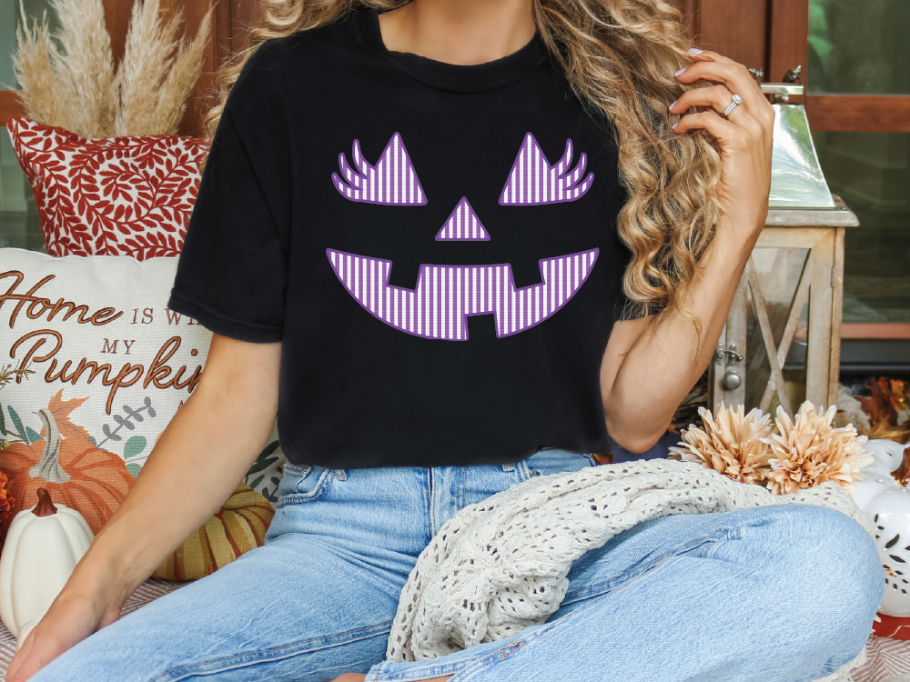 A woman wearing a black t-shirt with a purple jack-o-lantern graphic is sitting on a couch surrounded by fall decorations.
