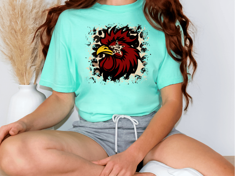A person wearing a turquoise t-shirt with a graphic of a rooster and a leopard print design, sitting on the floor.