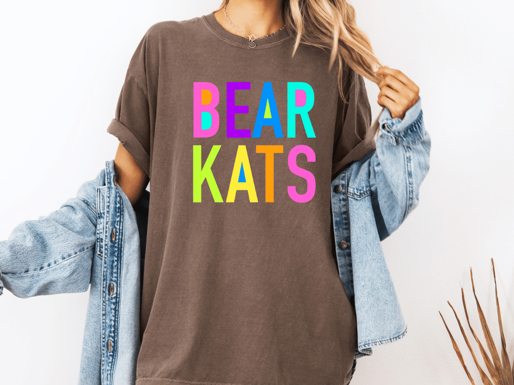 A person wearing a brown t-shirt with the text "BEAR KATS" in colorful letters, along with a denim jacket, stands against a white background.