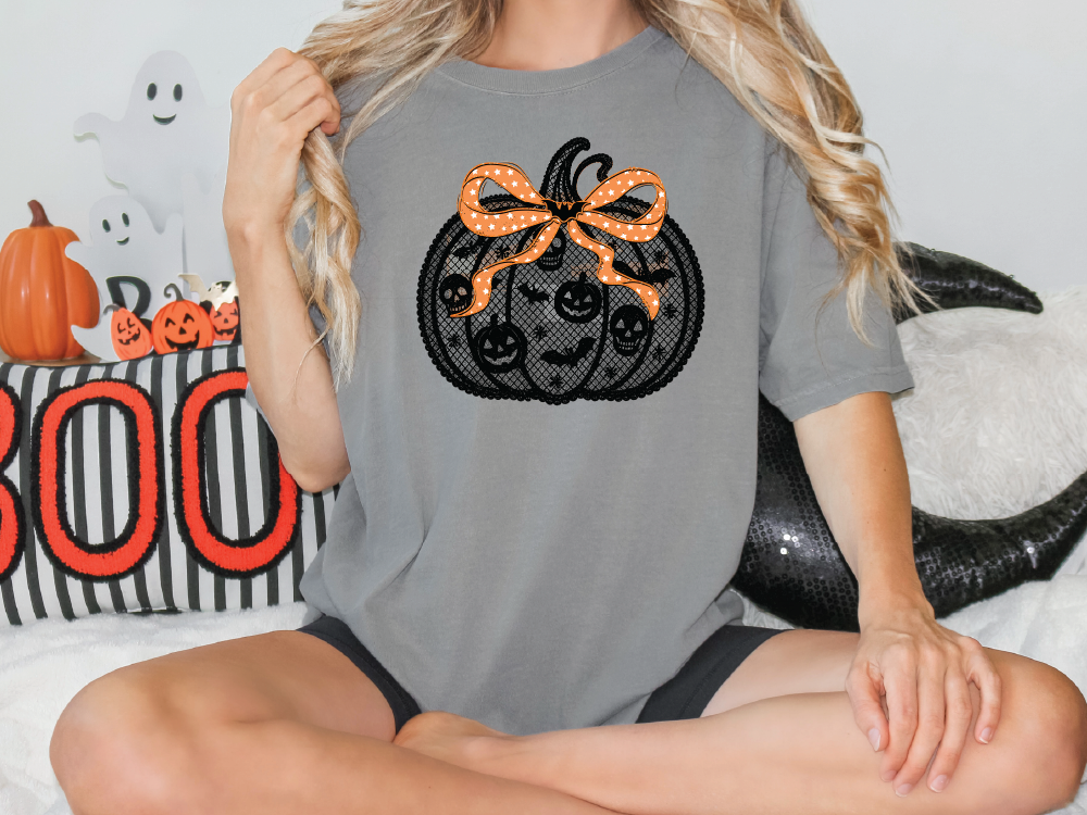 A woman is sitting cross-legged on a couch, wearing a gray t-shirt with a black pumpkin design and an orange ribbon bow.