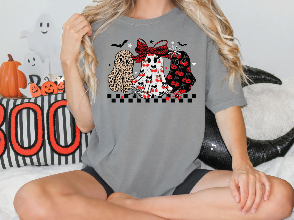 A woman wearing a gray t-shirt with a graphic of three ghostly figures and a bow on her head, sitting on a couch with Halloween decorations in the background.