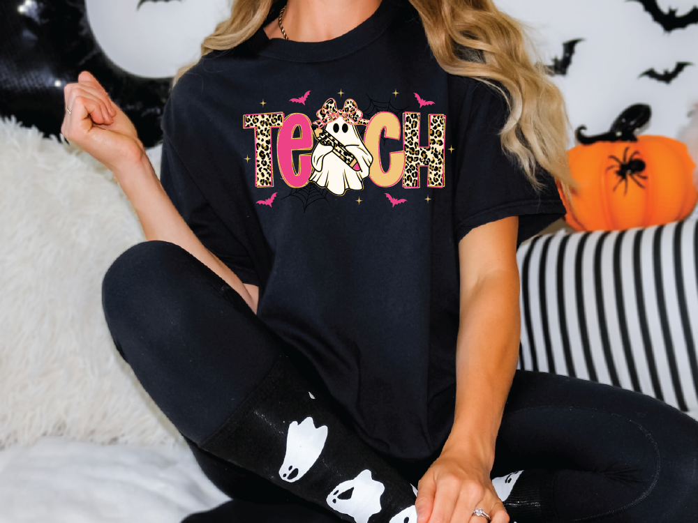 A person wearing a black t-shirt with a graphic design of a ghost and the word "Teach" on it, sitting on a couch with Halloween decorations in the background.
