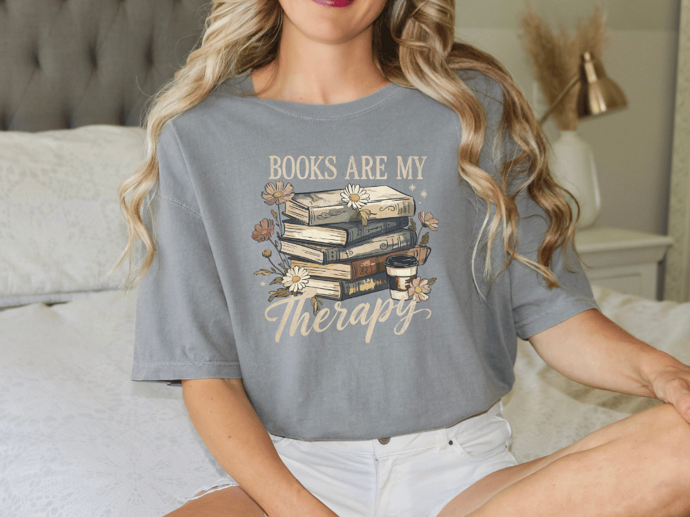 A woman wearing a gray t-shirt with a graphic design of books and flowers, along with the text "Books are my therapy".