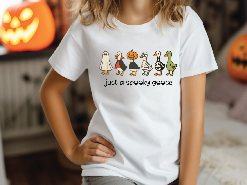 A person wearing a white t-shirt with a graphic design of five cartoon goose characters, including a ghost, a pumpkin, a skeleton, a mummy, and a green goose, along with the text "Just a spooky goose".
