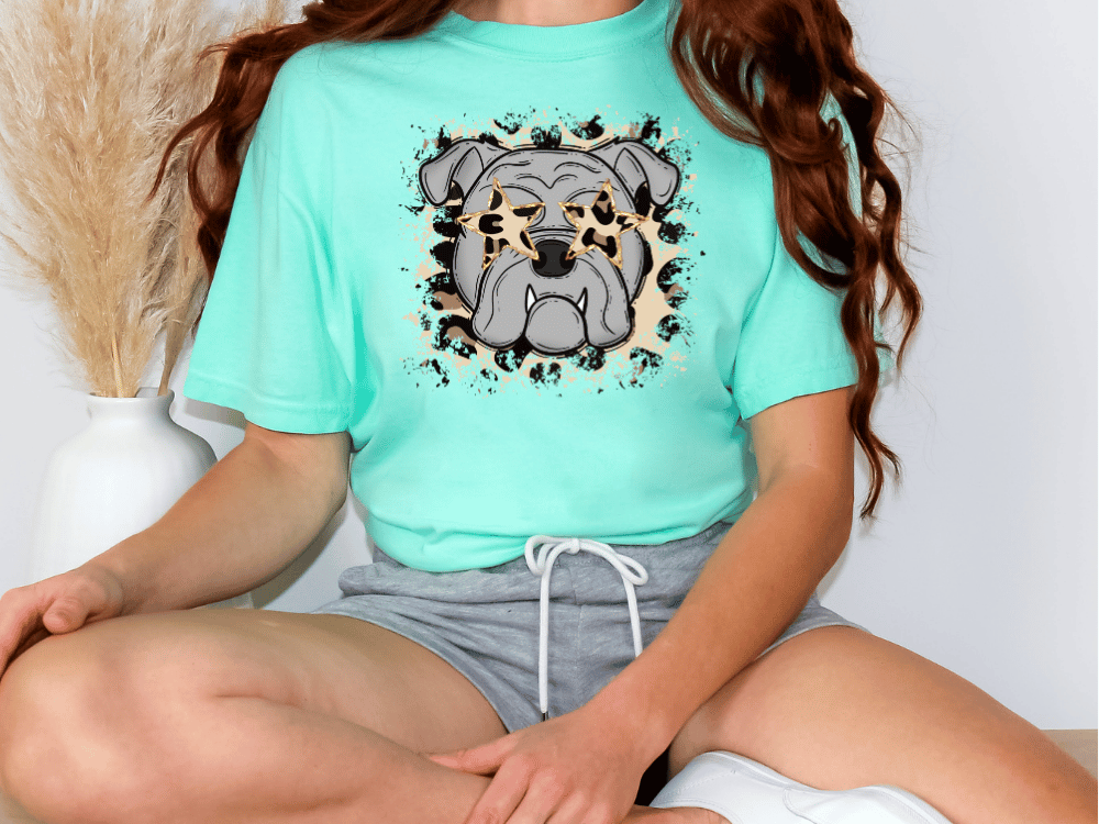 A person wearing a turquoise t-shirt with a graphic of a bulldog and a leopard print design, sitting on the floor with their legs crossed.