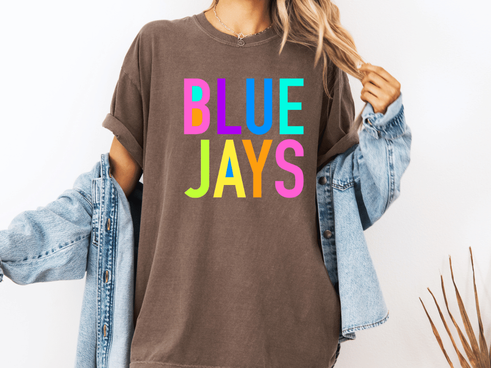 A person wearing a brown t-shirt with the text "BLUE JAYS" in colorful letters, along with a denim jacket, stands against a white background.