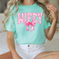 A woman wearing a mint green t-shirt with the word "WIFEY" and a pink bow on it, sitting on a bed.