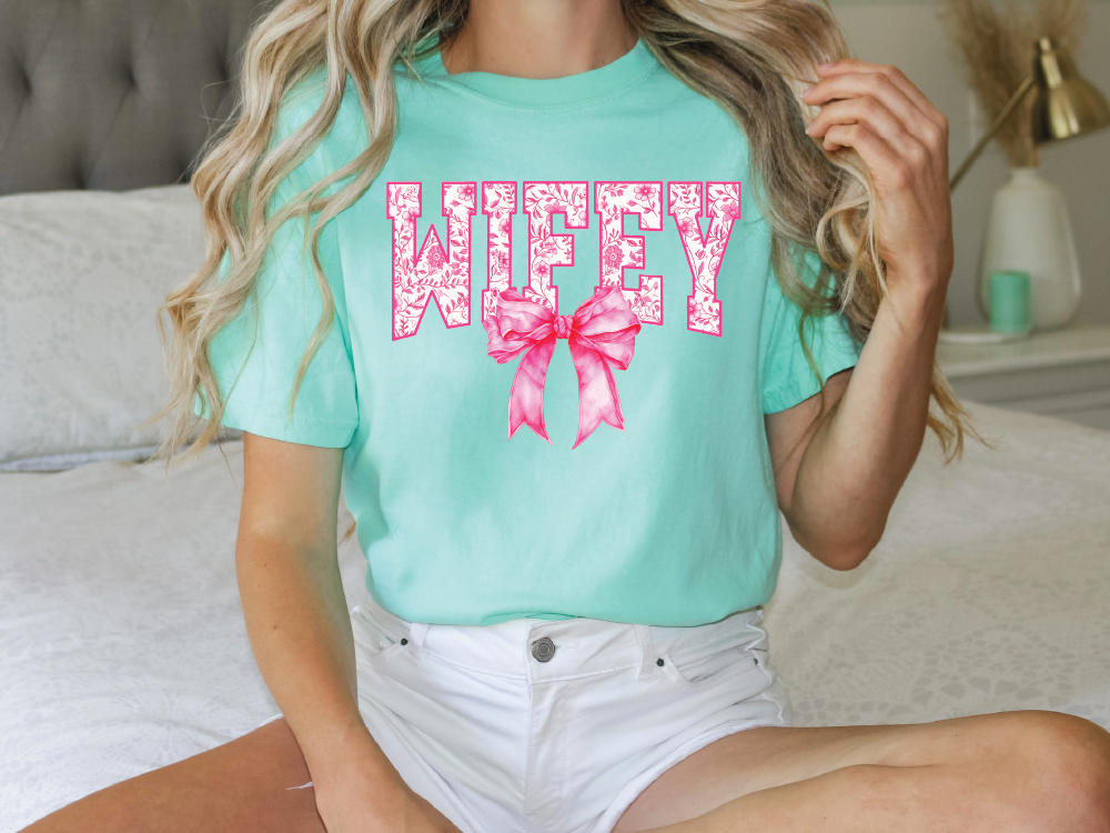 A woman wearing a mint green t-shirt with the word "WIFEY" and a pink bow on it, sitting on a bed.