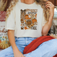 A woman wearing a white t-shirt with a graphic design of fall-themed items, including a football, pumpkins, and a cat, sitting on a couch with a red blanket.