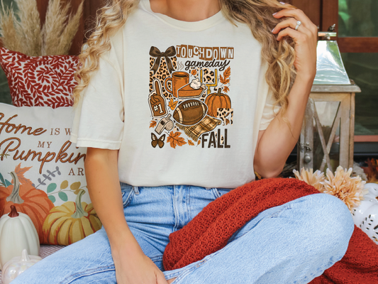 A woman wearing a white t-shirt with a graphic design of fall-themed items, including a football, pumpkins, and a cat, sitting on a couch with a red blanket.