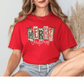 A woman wearing a red t-shirt with the word "MERRY CHRISTMAS" printed on it, sitting with her legs crossed and smiling.