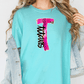 A woman wearing a turquoise t-shirt with a pink and black graphic design on it, and a denim jacket.