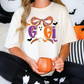 A woman wearing a white t-shirt with a Halloween-themed design is sitting on a couch, holding a pumpkin-shaped mug.