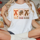A woman wearing a white t-shirt with a graphic design of a football and the text "TOUGH DOWN SEASON" is sitting on a bed with autumn-themed pillows.