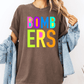 A person wearing a brown t-shirt with the text "BOMBERS" in colorful letters, along with a denim jacket, stands against a white background.