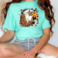 A woman wearing a turquoise t-shirt with a graphic of a leopard print eagle on it, sitting on the floor.