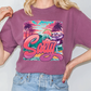 A person wearing a purple t-shirt with a graphic design featuring a beach scene, palm trees, and a surfboard.