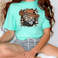 A woman wearing a turquoise t-shirt with a graphic of a jaguar wearing a cowboy hat, sitting on a chair.