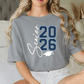 A woman wearing a grey t-shirt with the text "Graduation 26" printed on it, sitting on a bed.